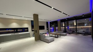 A modern living room with a sofa, ottoman, and a coffee table. There is a TV mounted on the wall, and a large window provides a view of the outside. - La Mirage Group Villas (Sukth)