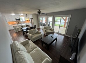 A spacious living area with a sofa, chairs, a coffee table, and a TV. There is a kitchen in the background with white cabinets and a dining area with a table and chairs.