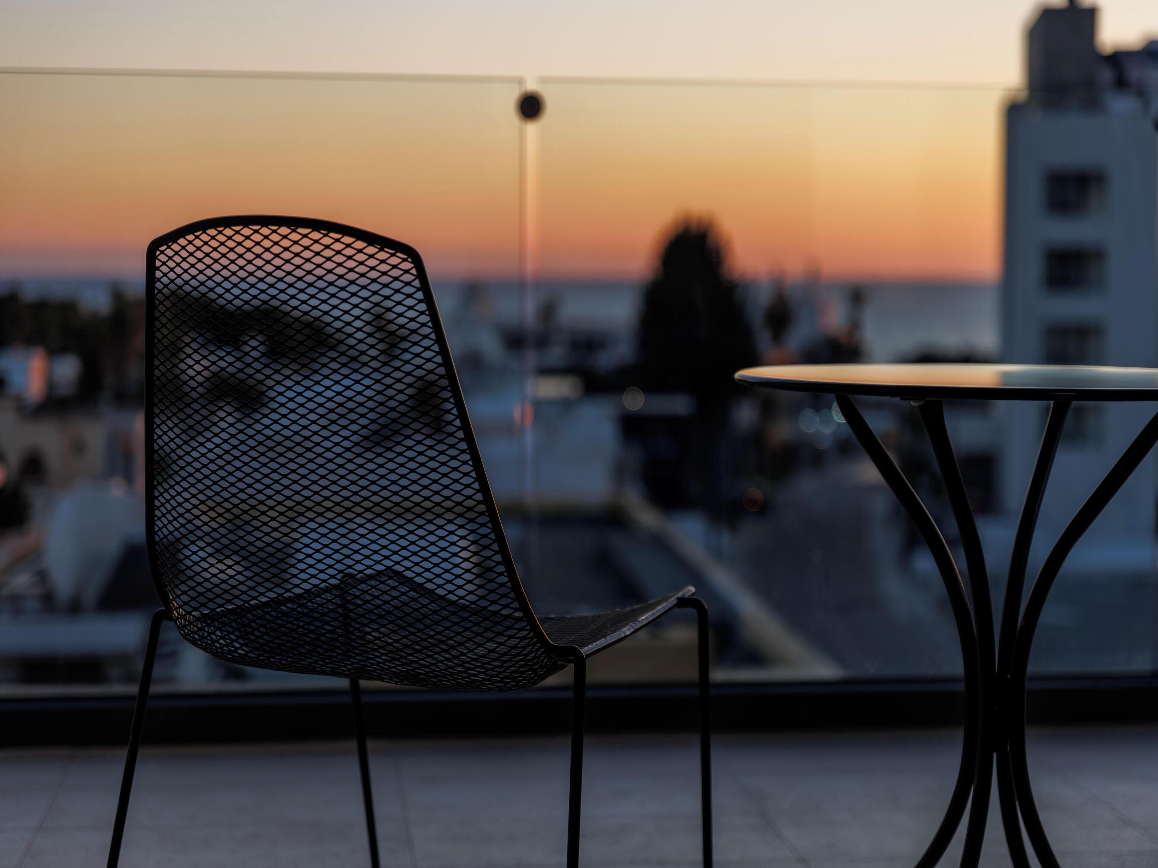 A modern chair with a mesh backrest and a round table with a black metal frame, positioned on a balcony overlooking a cityscape during sunset.