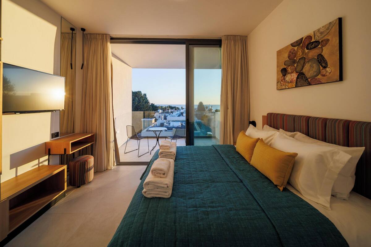 Una habitación de hotel moderna con una cama grande, un televisor, un balcón con vistas y un cuadro en la pared.