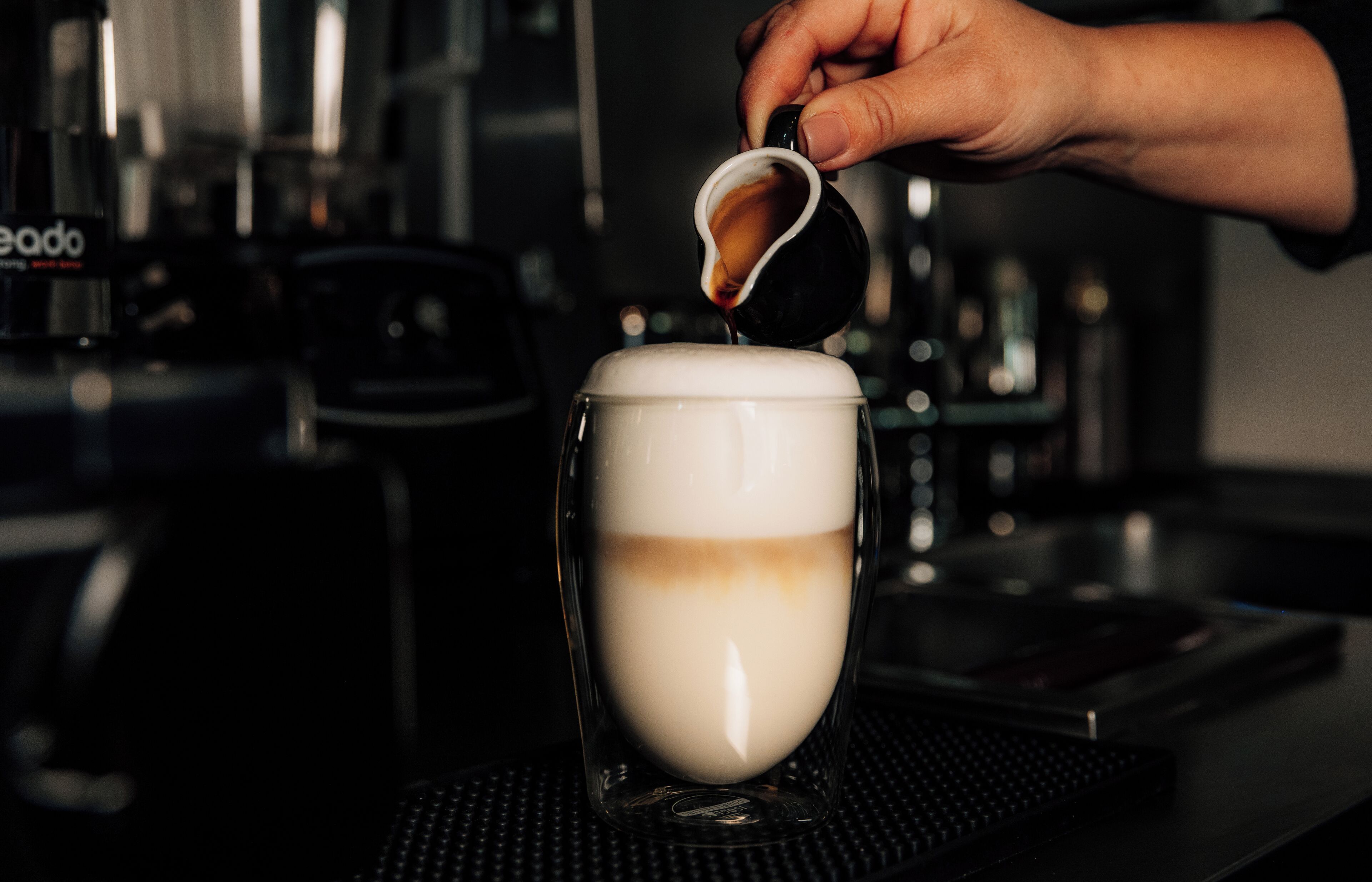 A hand pouring espresso into a cup, with a coffee machine in the background.