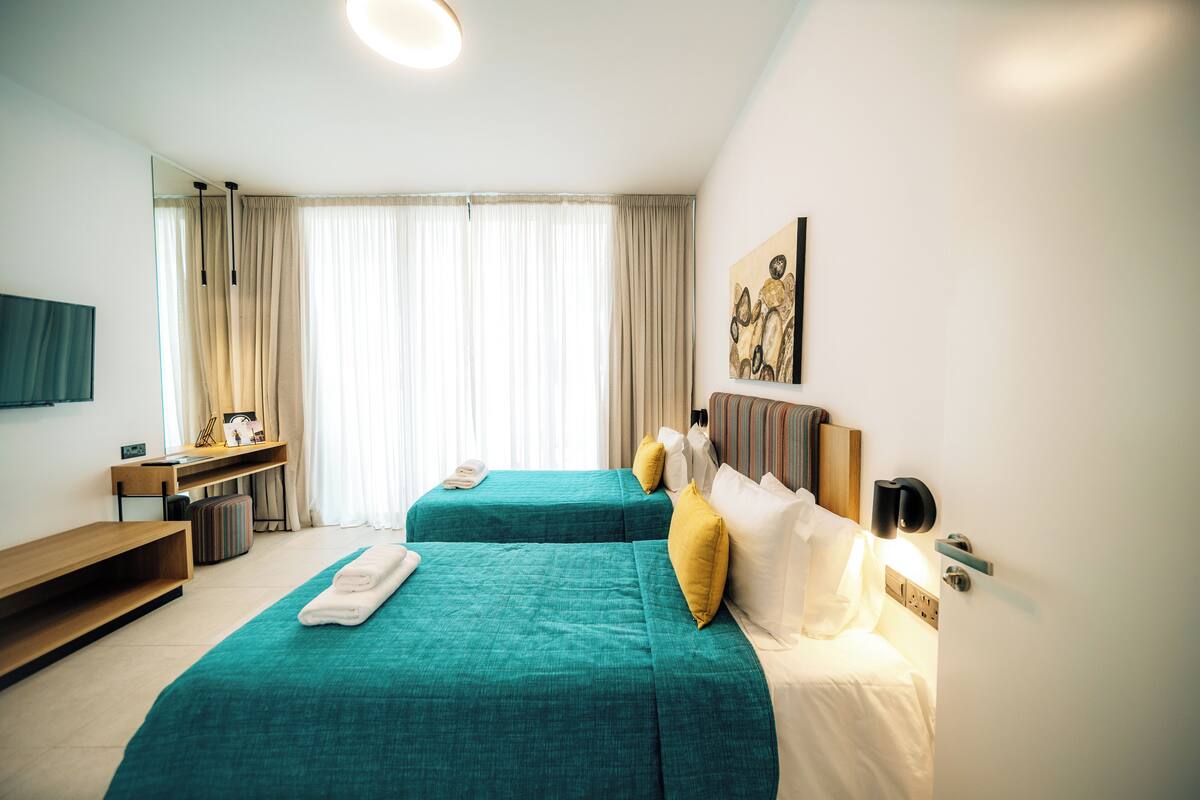 Habitación de hotel con dos camas, un escritorio, una silla, un televisor y un ventanal con cortinas.