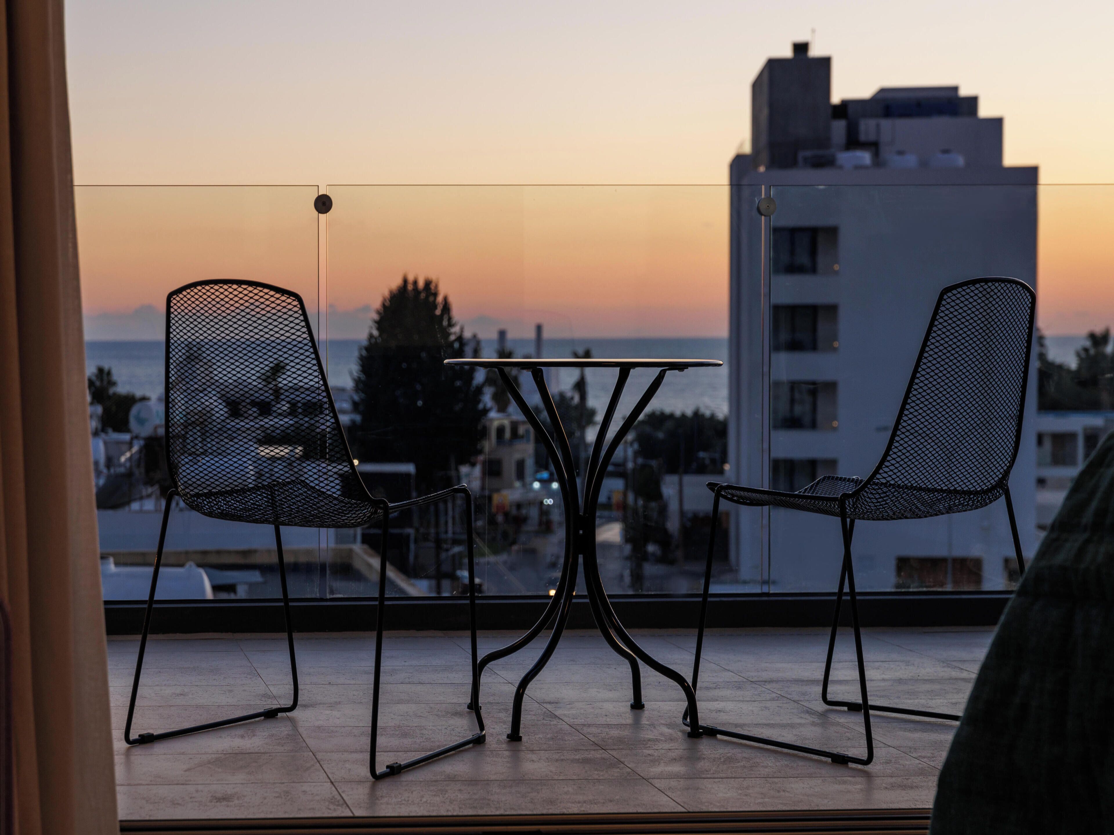Un balcón con dos sillas de malla y una mesita, con vistas a un paisaje urbano y al mar al atardecer.