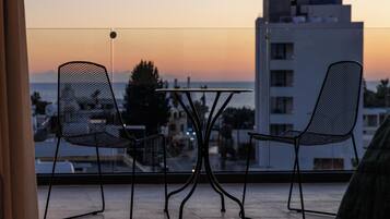 Un balcón con dos sillas de malla y una mesita, con vistas a un paisaje urbano y al mar al atardecer.