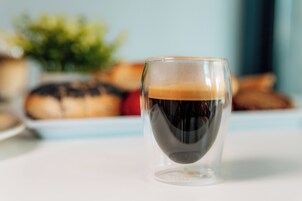 Una taza de café transparente con espuma sobre una superficie blanca.