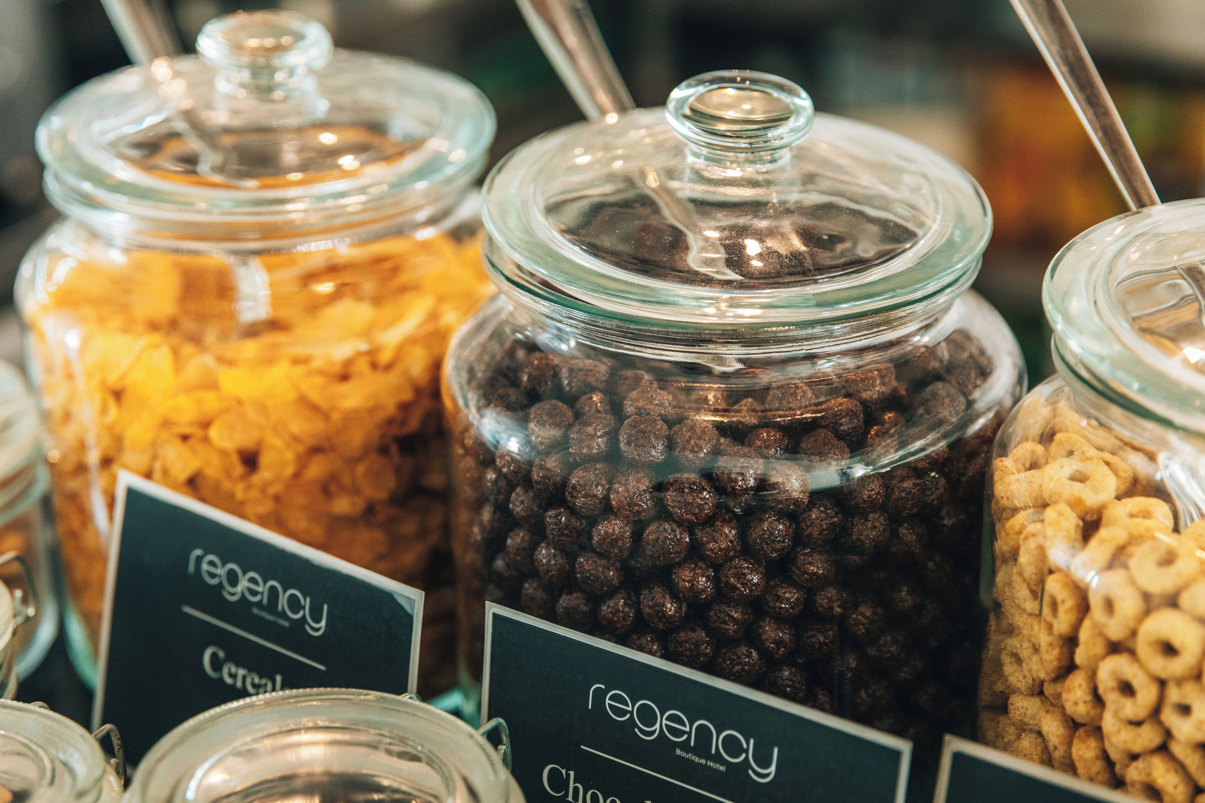 Une exposition de différents produits céréaliers présentés dans des bocaux en verre, chacun étant muni d’une étiquette indiquant, par exemple, « régence » ou « céréales ».