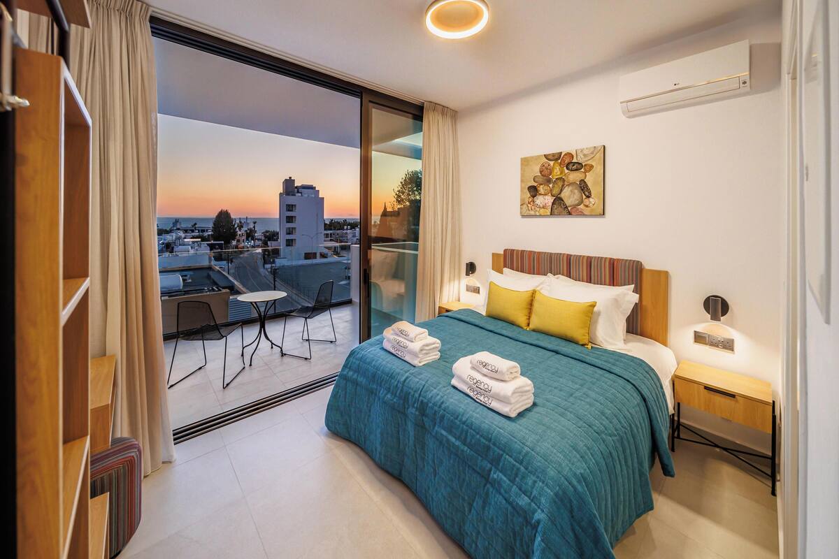 Una habitación de hotel moderna con balcón, cama con cubrecama color turquesa, mesita de noche y un cuadro en la pared.
