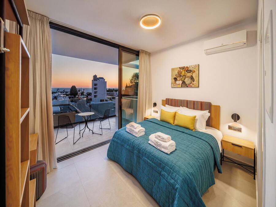 Una habitación de hotel moderna con balcón, cama con cubrecama color turquesa, mesita de noche y un cuadro en la pared.