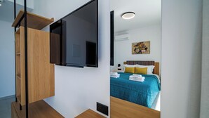 A modern bedroom with a large flat-screen TV mounted on the wall, a bed with a teal bedspread, and a wooden dresser with a mirror. - Regency  Boutique  Hotel (Paphos)