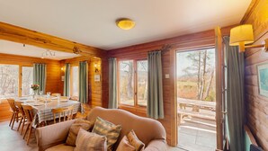 A cozy cabin interior with a dining area, a sofa, and large windows offering a view of the outdoors. - Balbeag Cottage - Pet friendly Log Cabin (Insh)