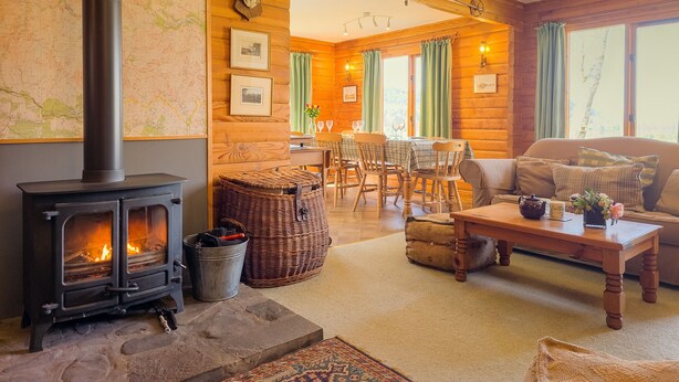 A cozy log cabin interior with a wood-burning stove, a dining area, and a sofa. - Balbeag Cottage - Pet friendly Log Cabin (Insh)