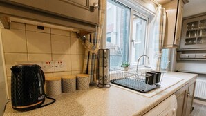 A kitchen with a countertop, a kettle, a coffee maker, a sink, and a window with curtains. - Cumbria House: Stunning Views, 3 Bedrooms & Garden (Fletchertown)