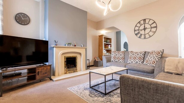 A living room with a flat-screen TV, a fireplace, a sofa with patterned cushions, a coffee table, and two wall clocks. - Cumbria House: Stunning Views, 3 Bedrooms & Garden (Fletchertown)