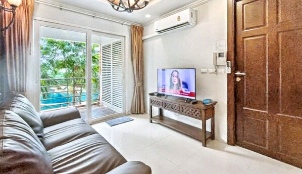 A living room with a sofa, a TV on a wooden stand, a sliding glass door to a pool, and an air conditioning unit on the wall.