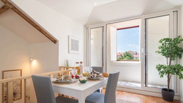 Un coin repas avec une table blanche dressée pour quatre personnes, une plante en pot et une vue à travers une porte-fenêtre coulissante.