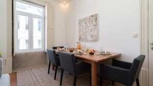 A dining area with a wooden table set for four, surrounded by chairs, a large abstract painting on the wall, and a window with blinds. - Modern Comfy Flat by HostWise (Porto)