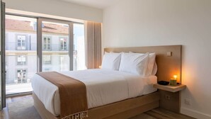 Habitación de hotel con una cama grande, una mesita de noche con lámpara y vistas a edificios a través de un ventanal.