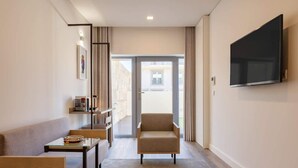 Una habitación de hotel moderna con sofá, una mesita, una silla, un televisor montado en la pared y un balcón con vistas.
