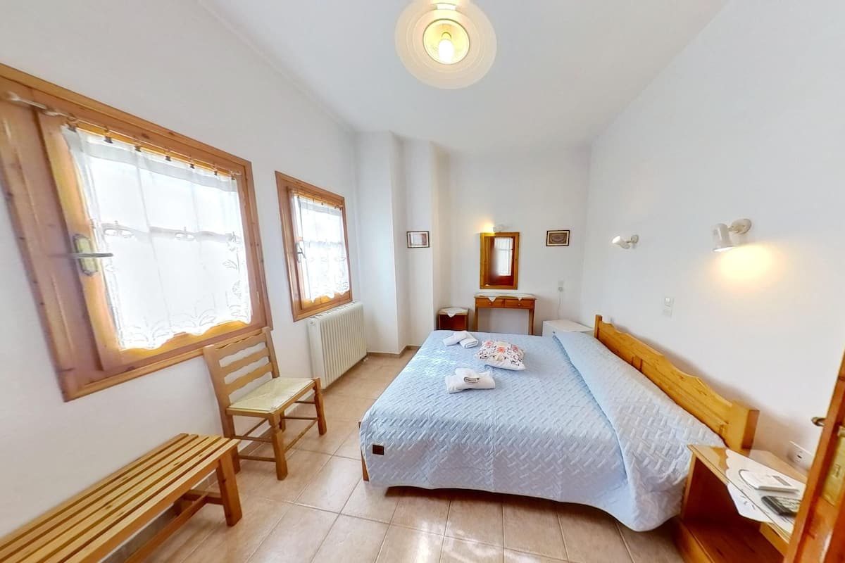 A bedroom with a bed, a wooden bench, a chair, a bedside table, a mirror, and a radiator.