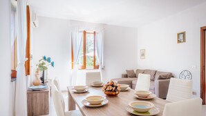 A dining area with a wooden table set for four, white chairs, and a sofa in the background. - Domus Antonini (Massa Lubrense)