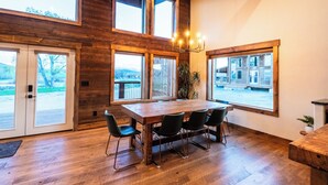 A spacious dining area with a large wooden table and chairs, large windows with a view of the outdoors, and a chandelier. - Luxury cabin near Cody: 2 kings, kids bunkroom, fire pit, country charm with easy access. (Cody)