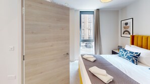 Habitación de hotel con puerta de madera, cama con ropa de cama estampada, mesita de noche, cuadro enmarcado en la pared y vistas a edificios.
