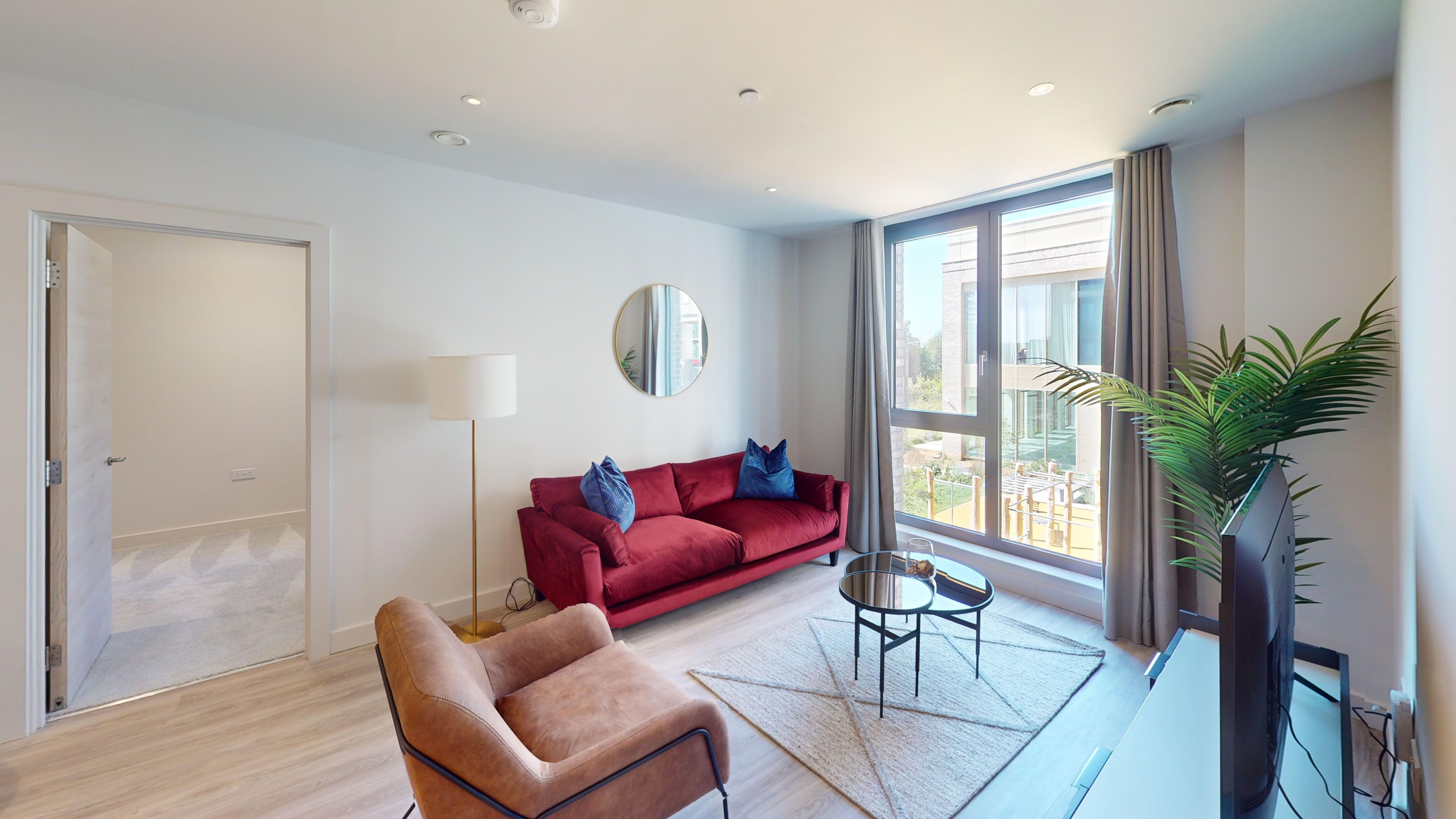 Un salon moderne avec un canapé rouge, un fauteuil marron, un miroir rond, une lampe sur pied, une petite table avec un vase et une grande fenêtre donnant sur des bâtiments et de la verdure.