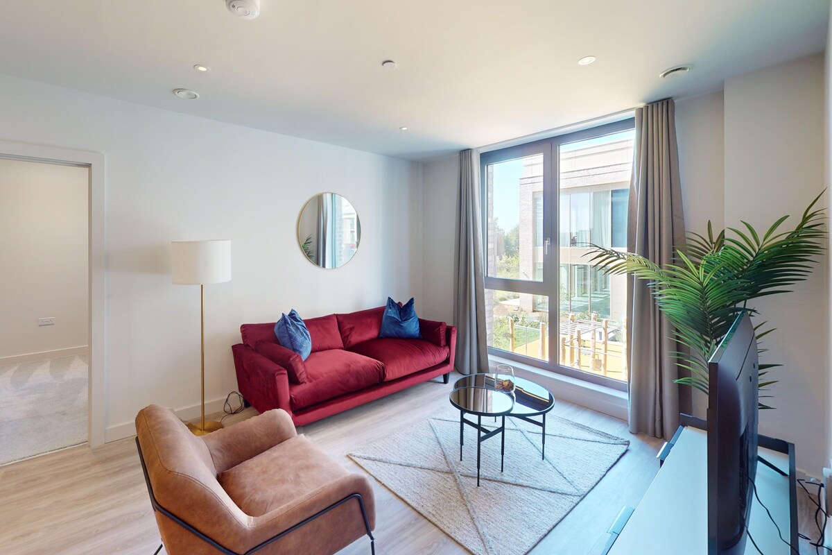 A modern living room with a red sofa, a brown armchair, a round mirror, a floor lamp, a small table with a glass, and a large window with a view of buildings and greenery.