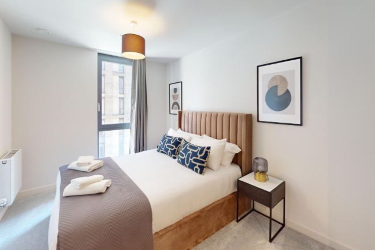 A modern hotel room with a large bed, a bedside table with a lamp, and a window with curtains.