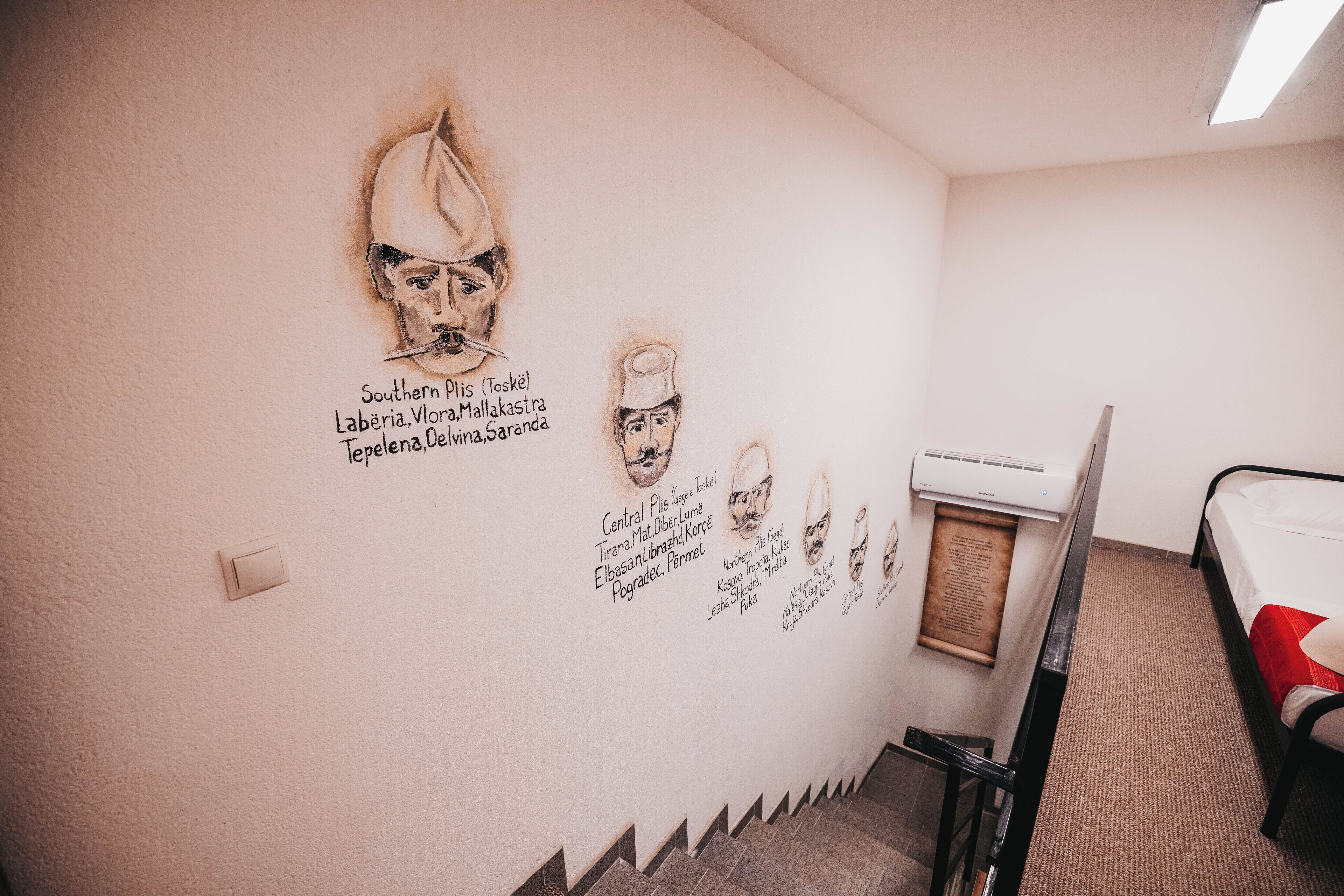 A room with a wall decorated with portraits and names, a bed, a radiator, and a small table.