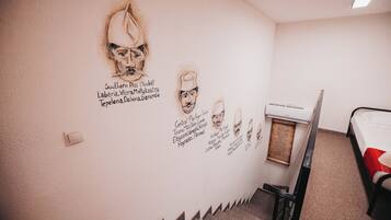 A room with a wall decorated with portraits and names, a bed, a radiator, and a small table.
