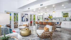 A modern open-plan living and dining area with a kitchen, wicker furniture, and a large window overlooking greenery. - Serenity Oasis~Pool!~Putt-Putt!~Kayaks~Bikes~Spa (Marathon)