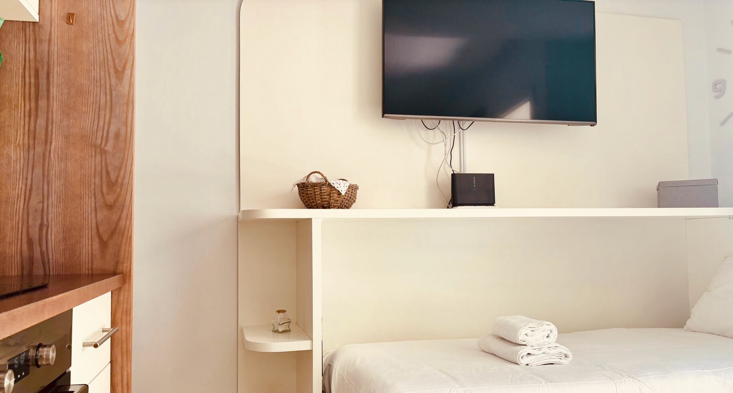 Ein Hotelzimmer mit Wand-Fernseher, einem Holzkasten, einer kleinen Ablage mit Korb und Dekoration sowie einem ordentlich bezogenen Bett mit gefalteten Handtüchern.