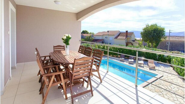 A balcony with a wooden table and chairs, a vase with flowers, and a view of a swimming pool and houses. - Villa Isabella (Krk)