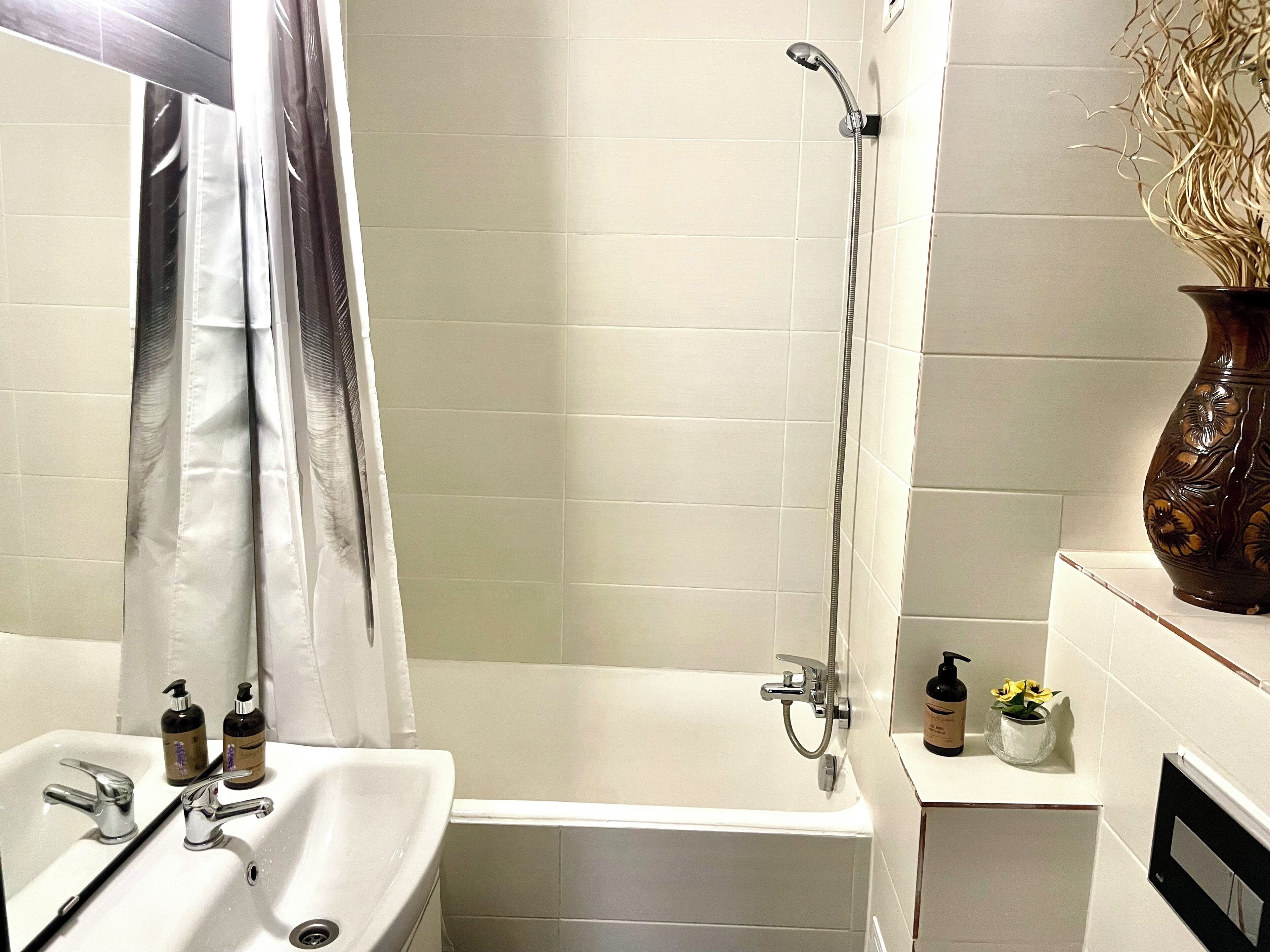 A bathroom with a white sink, mirror, shower, and a vase with dried flowers on a shelf.