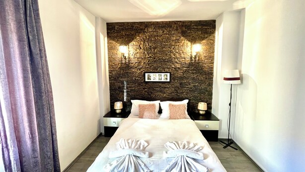 Habitación de hotel con pared de piedra, una cama con dos almohadas, dos mesitas de noche con lámparas y un cuadro en la pared.