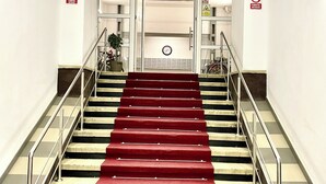 Una escalera con peldaños alfombrados de rojo y barandas de metal, que conduce a un espacio con puertas de vidrio y un reloj en la pared.