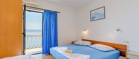 A bedroom with a bed, a nightstand, a window with blue curtains, and a view of the sea.