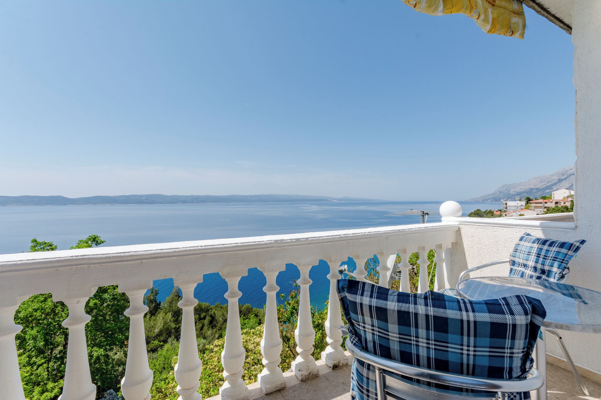 Ein Balkon mit weißer Brüstung, einem Tisch und einem Stuhl, mit Blick auf das Meer und ferne Berge.