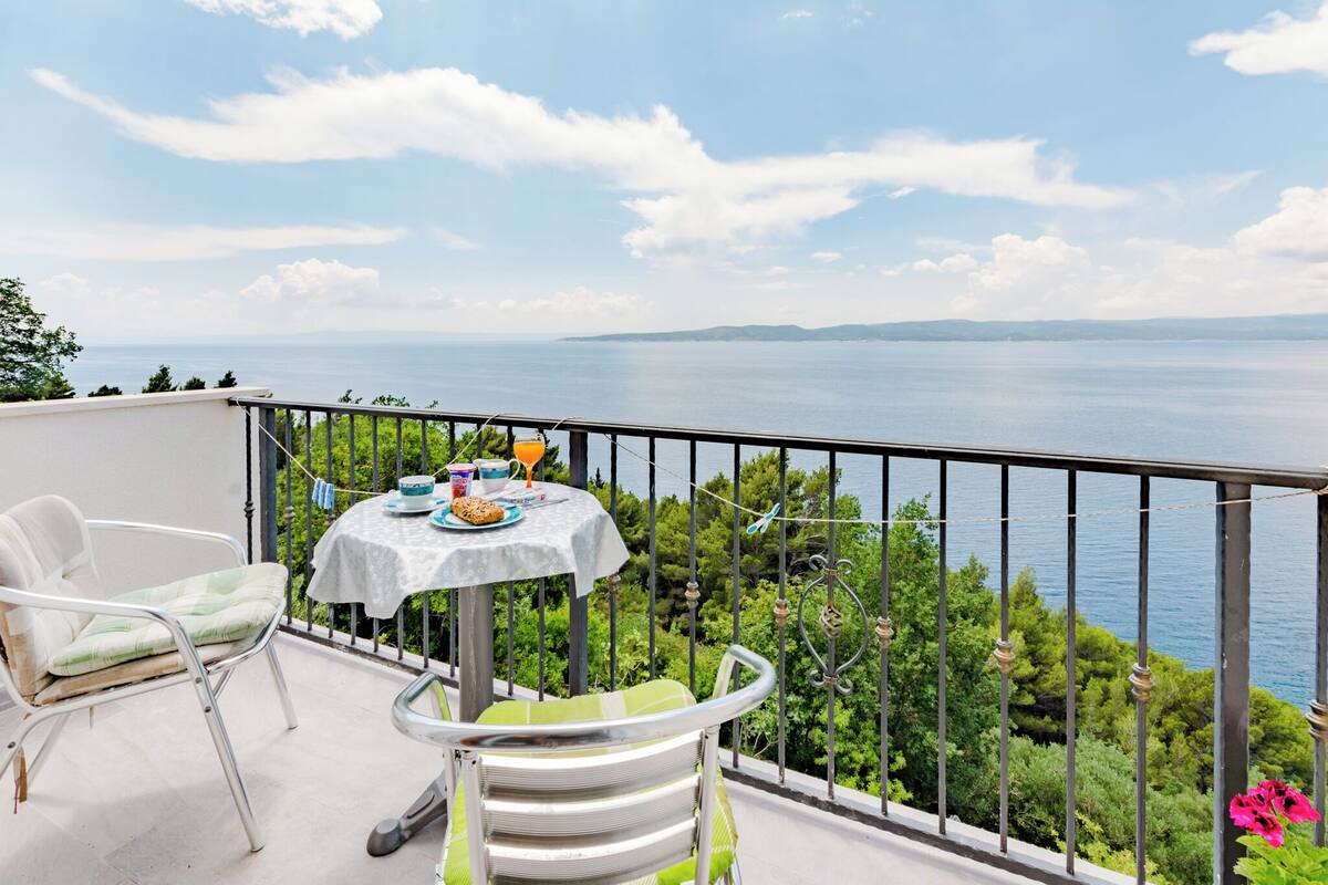 A balcony with a table set for breakfast, overlooking a scenic view of the sea and greenery.