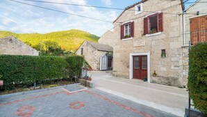 A stone building with a red door and window shutters, surrounded by greenery and a paved area with hexagonal tiles. - Villa Nives (Ston)
