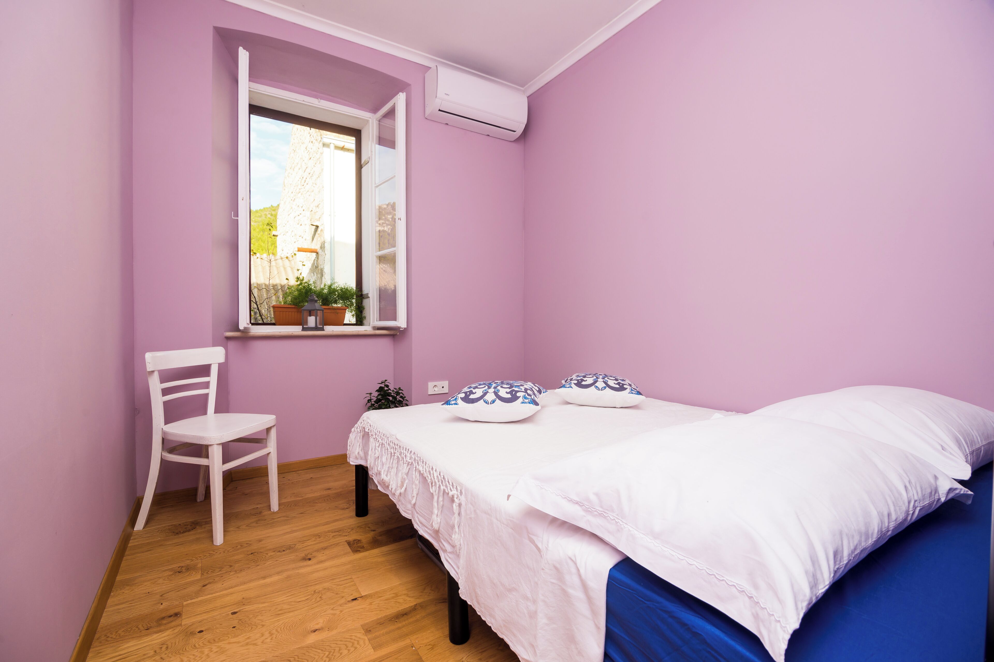 A bedroom with a bed, a chair, a window with a view, and an air conditioning unit.