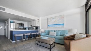 Et moderne køkken med en central ø, et opholdsområde med en blå sofa og en brun lænestol, samt et stort maleri af en strandscene.