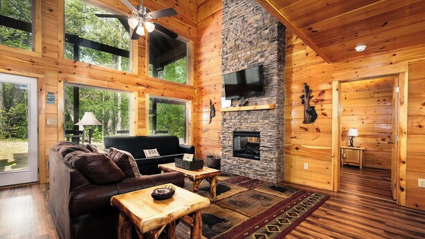 A wooden cabin interior with a stone fireplace, leather sofa, and a television mounted on the wall. - 2BR, sleeps up to 10, less than 8 minutes to Dollywood, Island, Parkway! (Pigeon Forge)