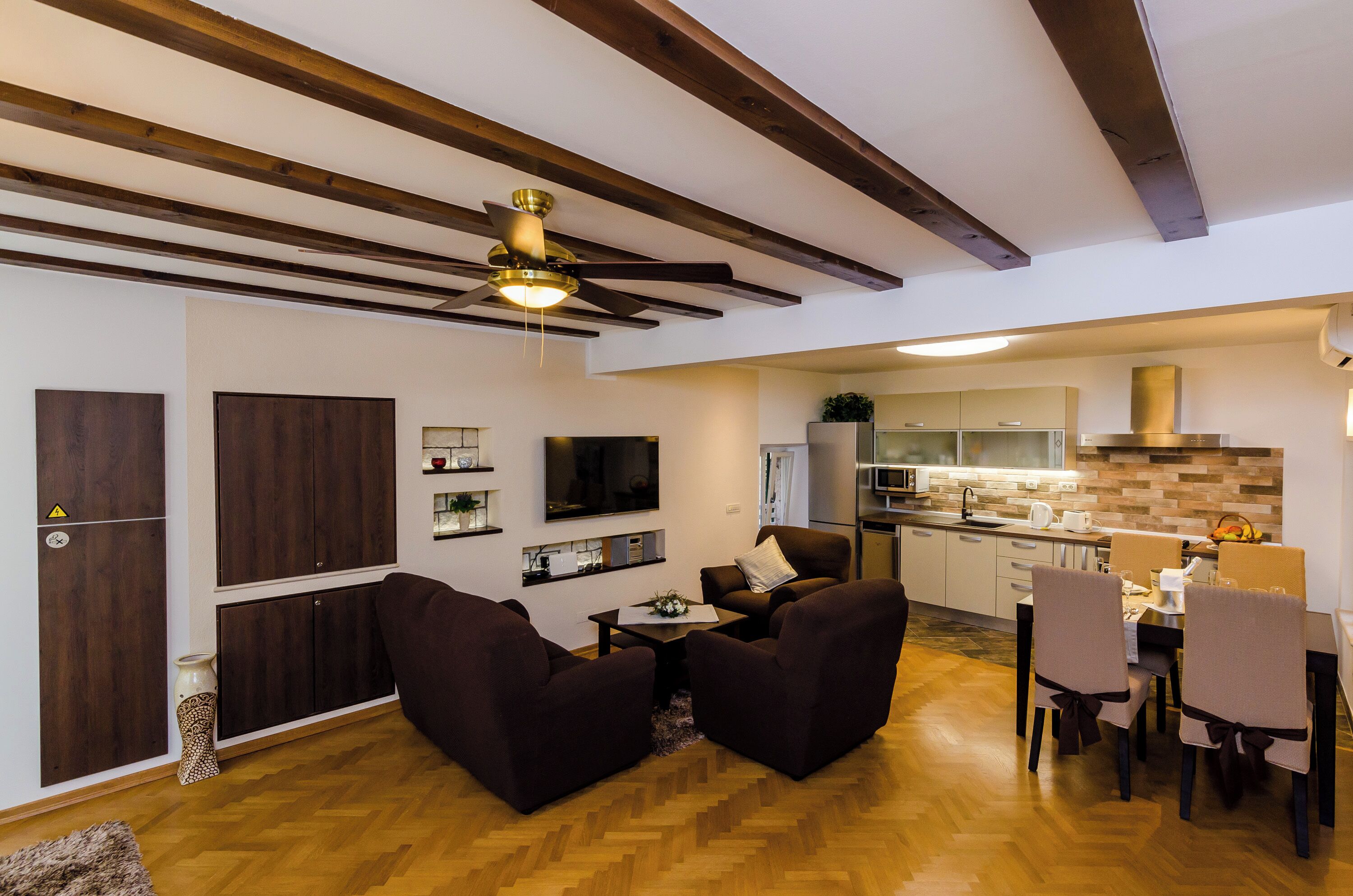 Una sala de estar moderna con sofás oscuros, una mesa de centro y un televisor. El cuarto tiene pisos de madera y un ventilador de techo.