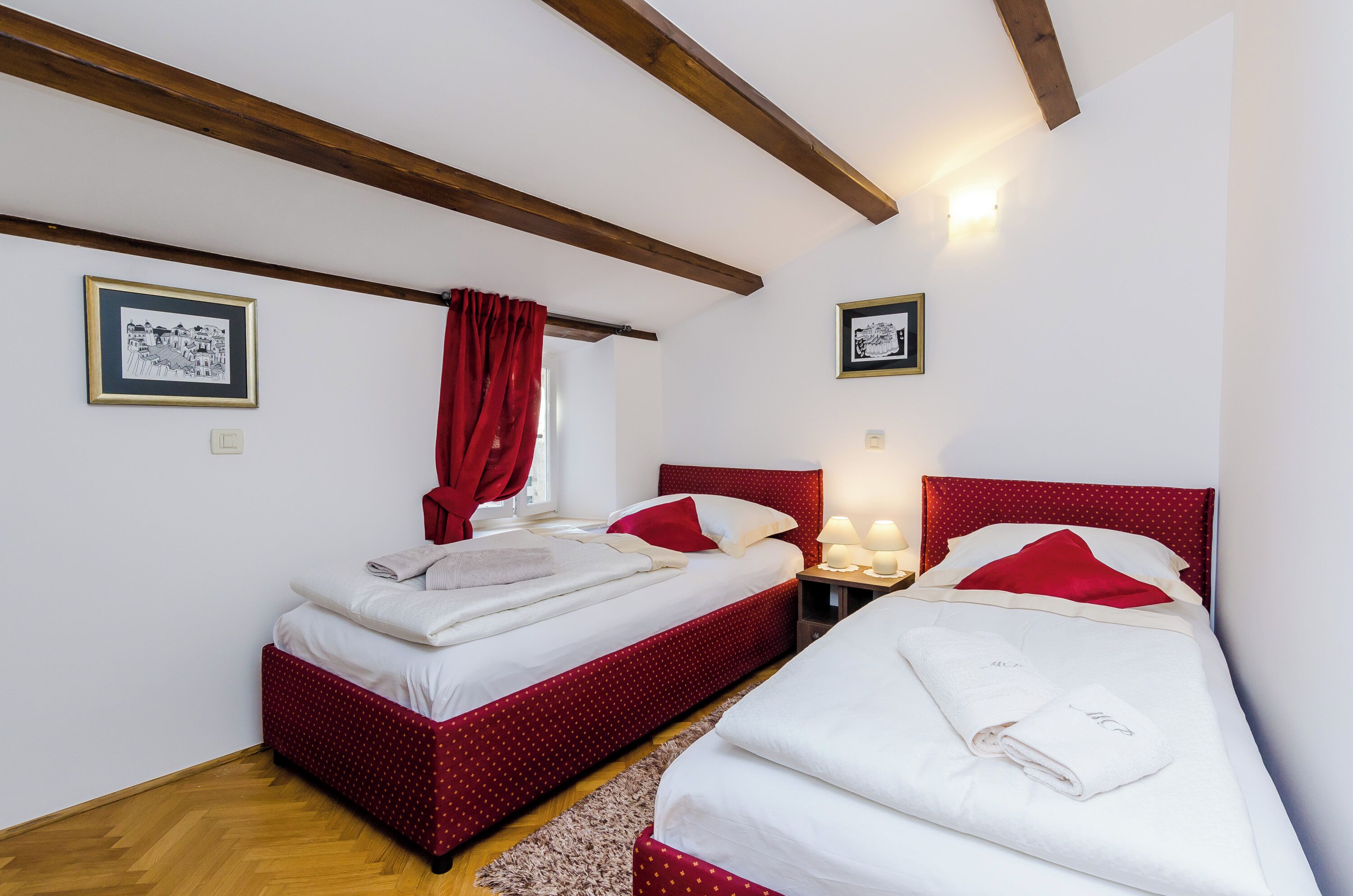 Een hotelkamer met twee bedden, rood-witte beddengoed, houten balken in het plafond en twee ingelijste foto's aan de wanden.