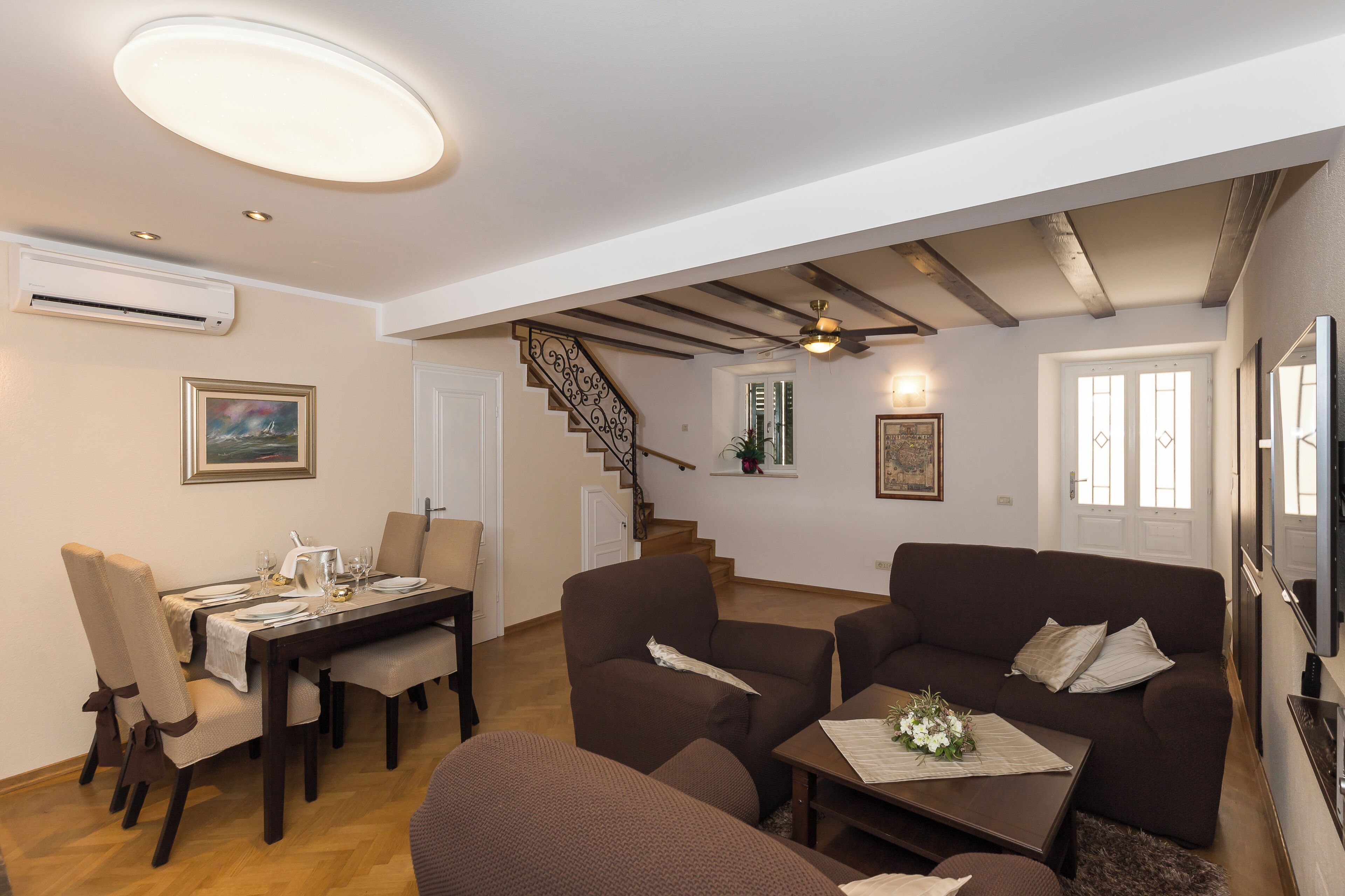 A living room with a brown sofa, a dining table set for four, and a staircase leading to another level.