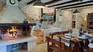 A rustic kitchen with a stone wall, wooden beams, a fireplace, and a dining area with a wooden table and benches.