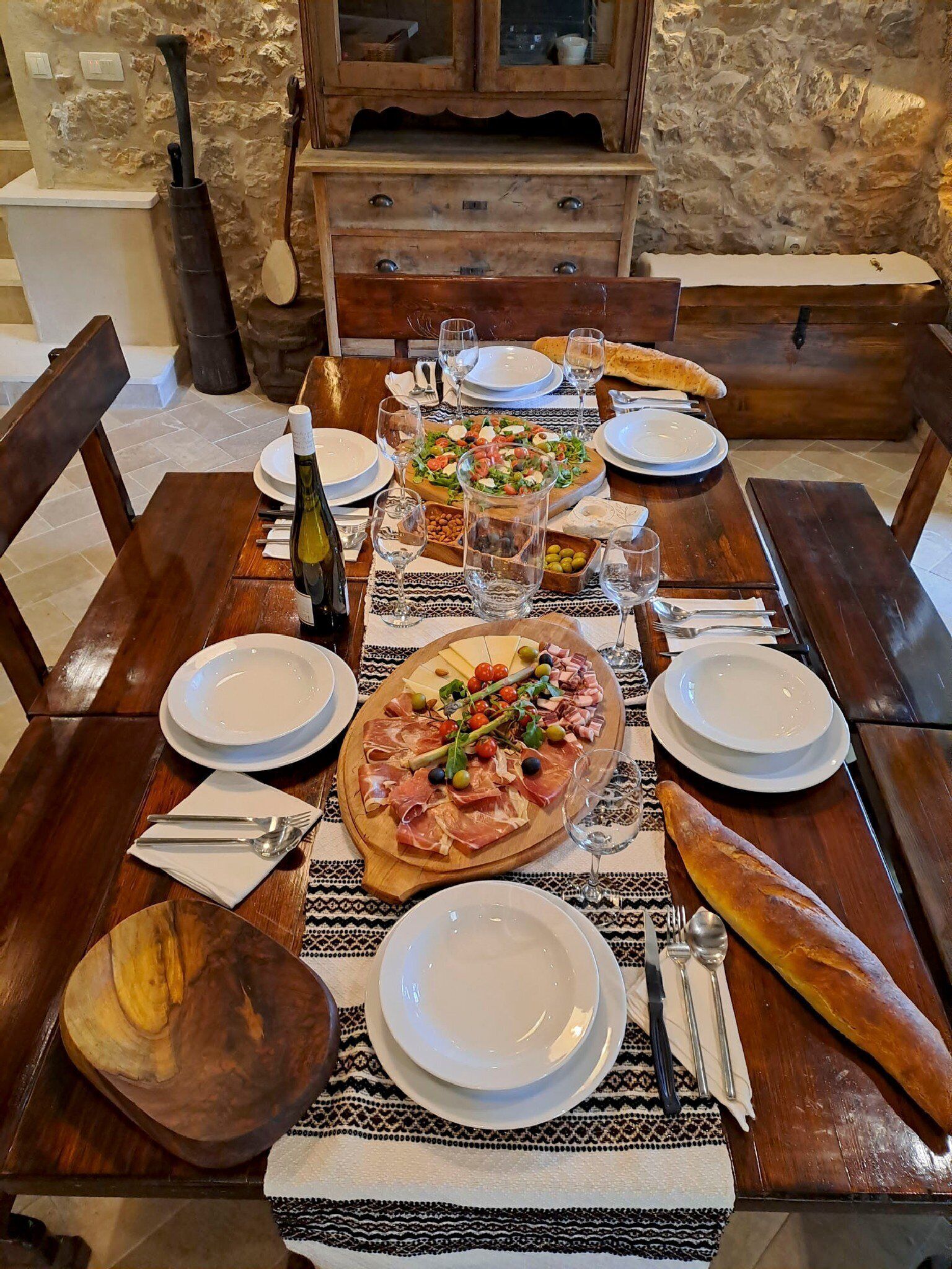 Ein Esstisch ist gedeckt mit Tellern, Besteck, einer Flasche, Gläsern und verschiedenen Speisen, darunter Pizza, eine Wurstplatte und Brot.