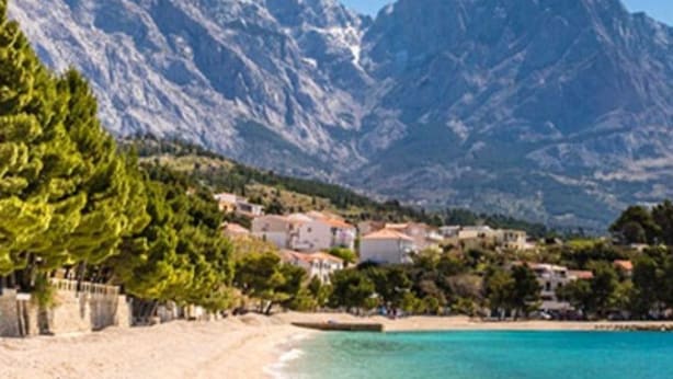 A beach with clear turquoise water, a sandy shore, and a backdrop of rocky mountains.